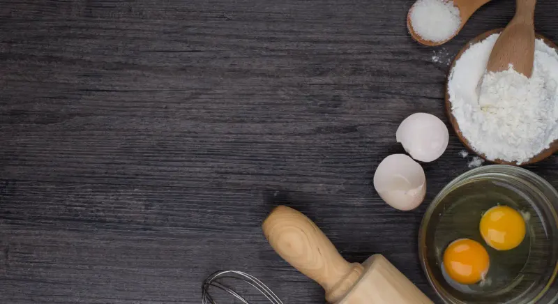 Eggs, flour, a whisk and a rolling pin on a dark wood counter, mid-recipe